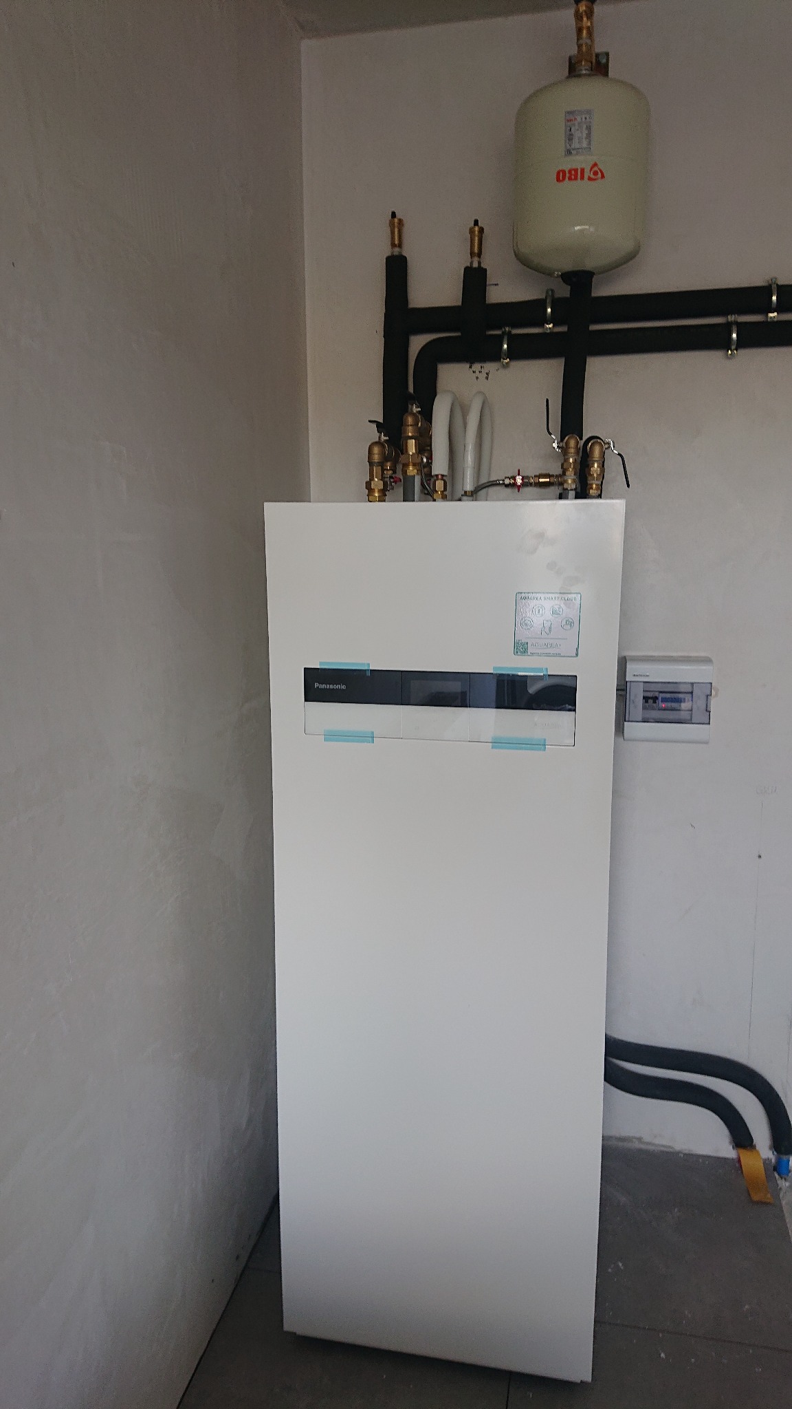 Biała pompa ciepła Panasonic zamontowana w pomieszczeniu, z widocznymi rurami instalacji i zbiornikiem wyrównawczym IBO na ścianie.