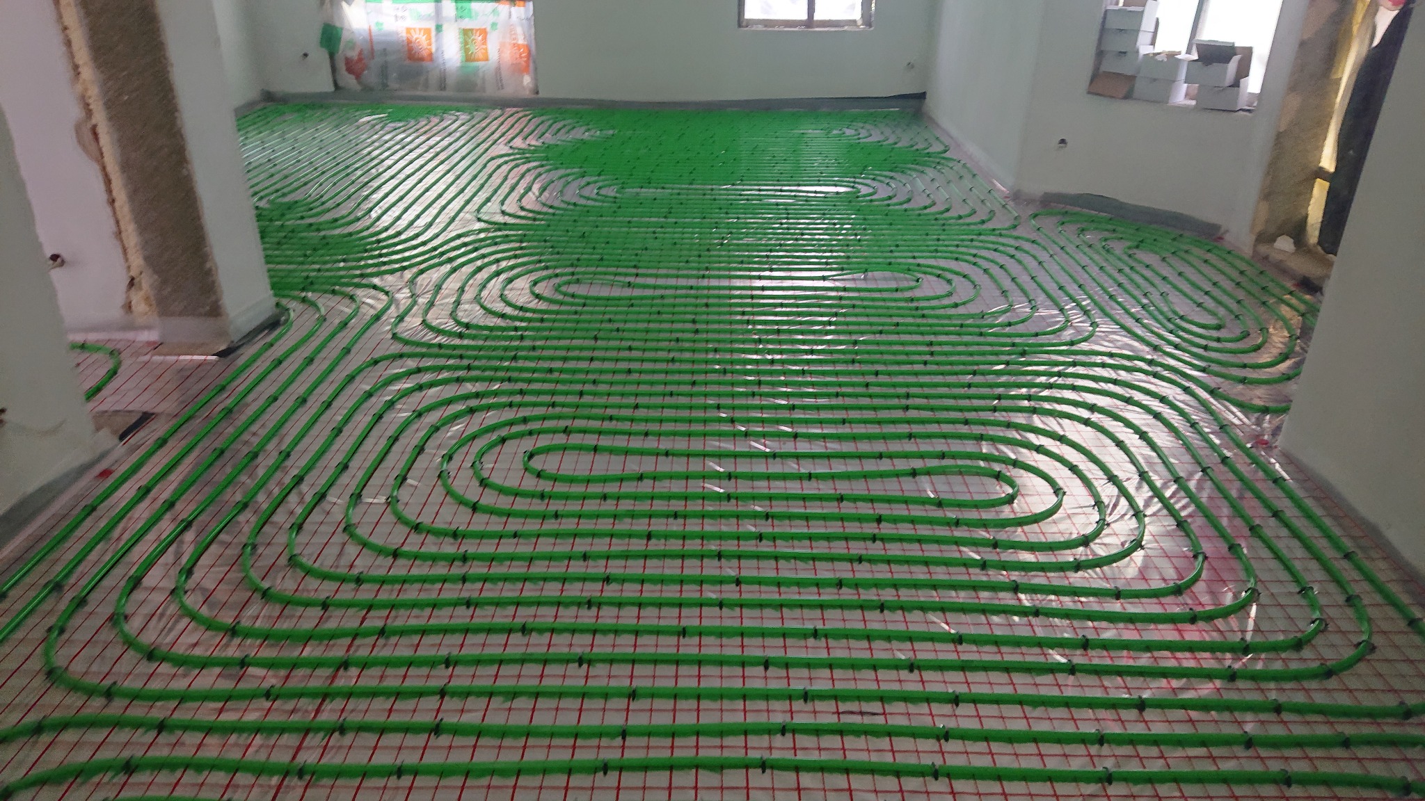 Zielone rury ogrzewania podłogowego ułożone na siatce z czerwonymi liniami w jasnym pomieszczeniu w trakcie montażu.