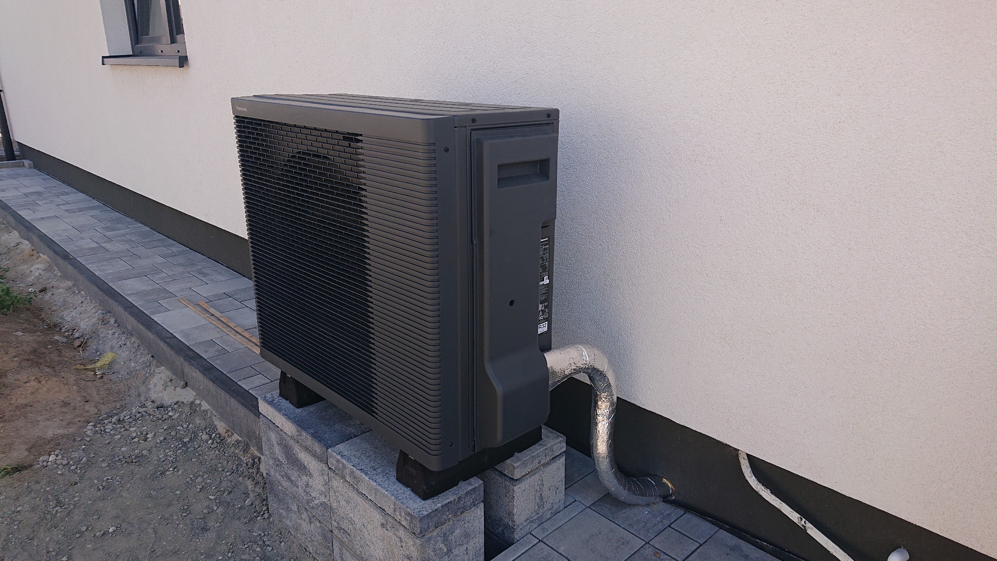 Czarna pompa ciepła powietrze-woda Panasonic Aquarea, zamontowana na betonowych bloczkach przy ścianie domu z ociepleniem, z widoczną izolowaną rurą odprowadzającą.
