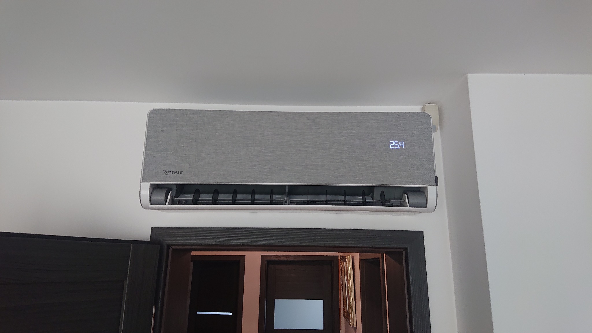 Szary klimatyzator Rotenso zamontowany na białej ścianie nad ciemnymi drzwiami, wyświetlający temperaturę 25.4 stopnia Celsjusza.