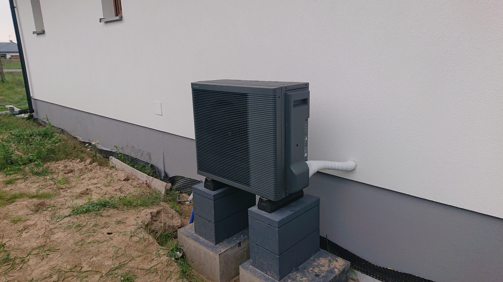 Szara pompa ciepła zamontowana na betonowych bloczkach przy ścianie budynku z widocznymi przyłączami.