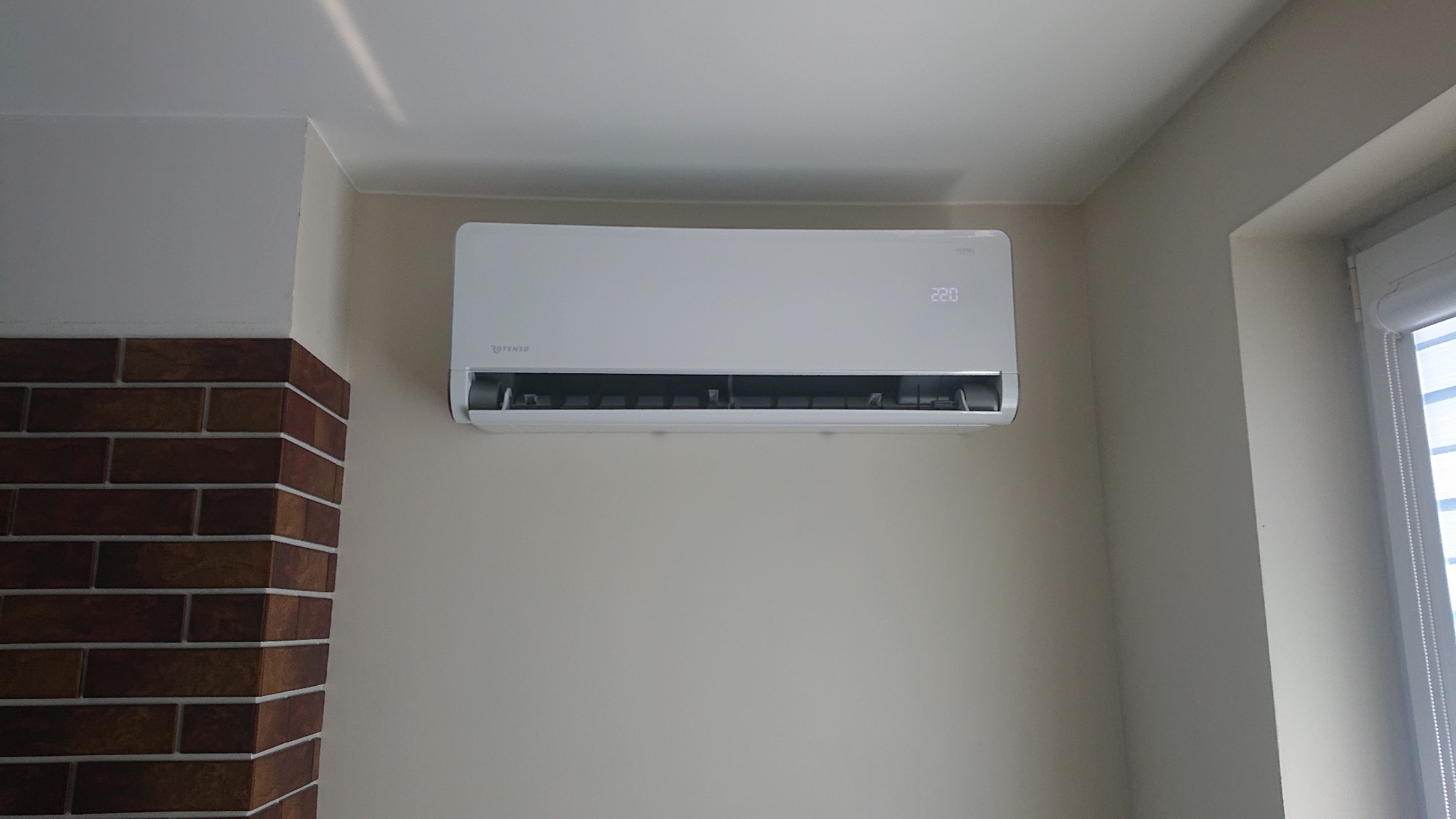 Biały klimatyzator Rotenso zamontowany na ścianie w pomieszczeniu w Zdunach, widoczny fragment ściany z cegły i okno z roletą.