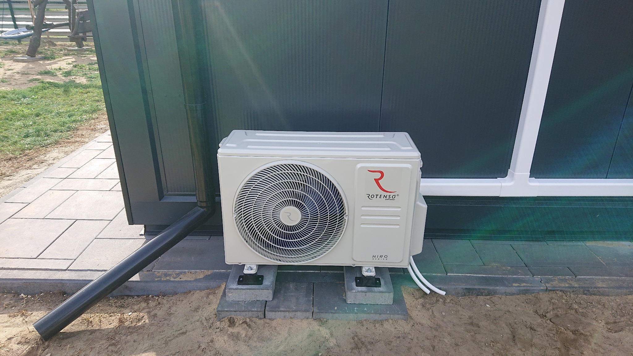Zewnętrzna jednostka klimatyzacji Rotenso Hiro Series zainstalowana na betonowych bloczkach, z widoczną rurą odpływową i fragmentem elewacji budynku.