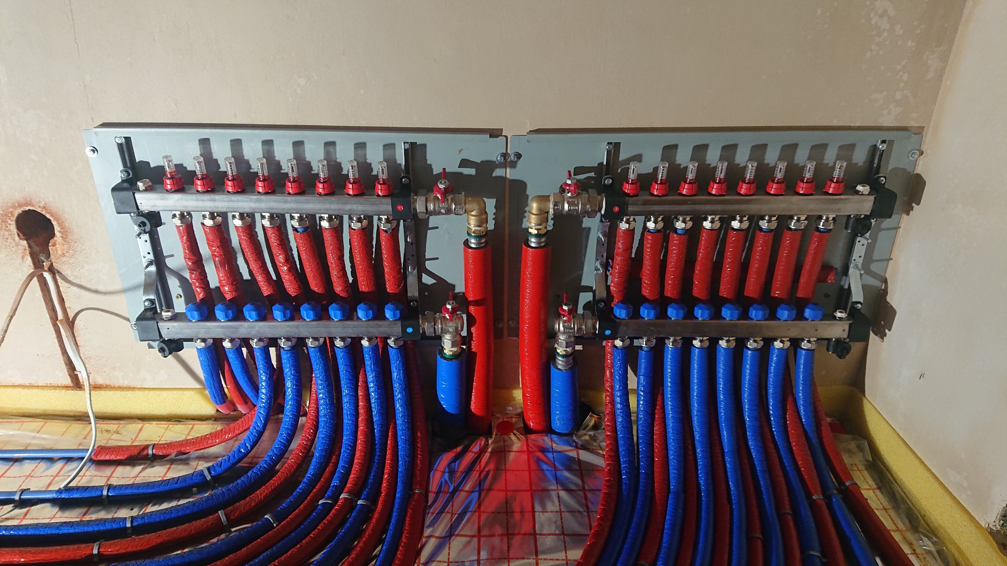 Rozdzielacz ogrzewania podłogowego z czerwonymi i niebieskimi rurami, zamontowany na ścianie w trakcie instalacji, widoczne manometry i zawory.