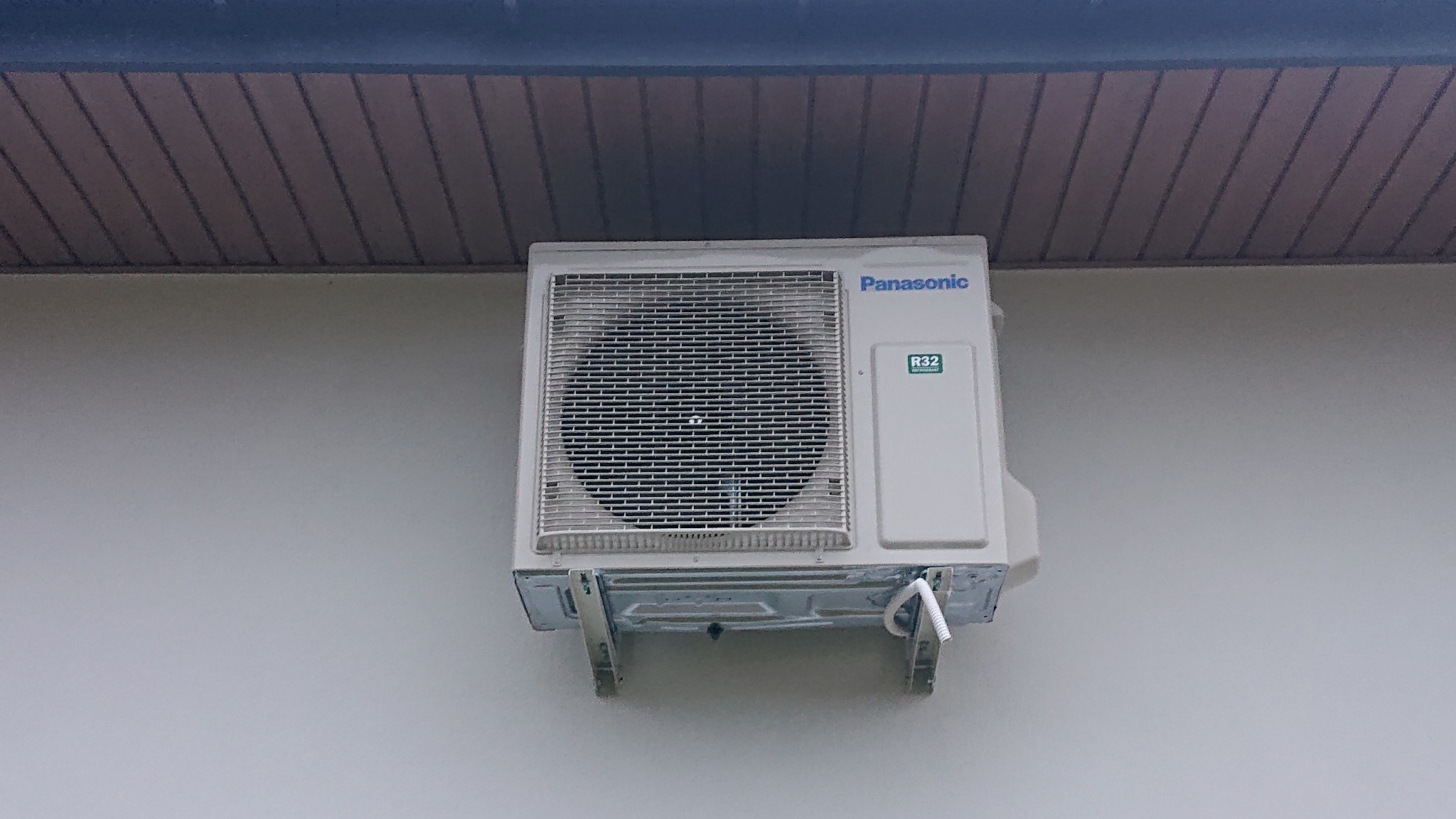 Zewnętrzna jednostka klimatyzacji Panasonic zamontowana na jasnej ścianie budynku pod okapem.