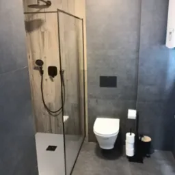 Nowoczesna łazienka z kabiną prysznicową typu walk-in z drewnopodobną ścianą, szarymi płytkami, podtynkową toaletą i czarnymi akcentami.