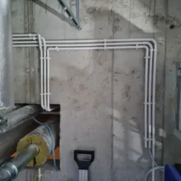 Białe rury poprowadzone wzdłuż surowej, betonowej ściany, widoczne przewody w izolacji i skrzynka narzędziowa na podłodze.