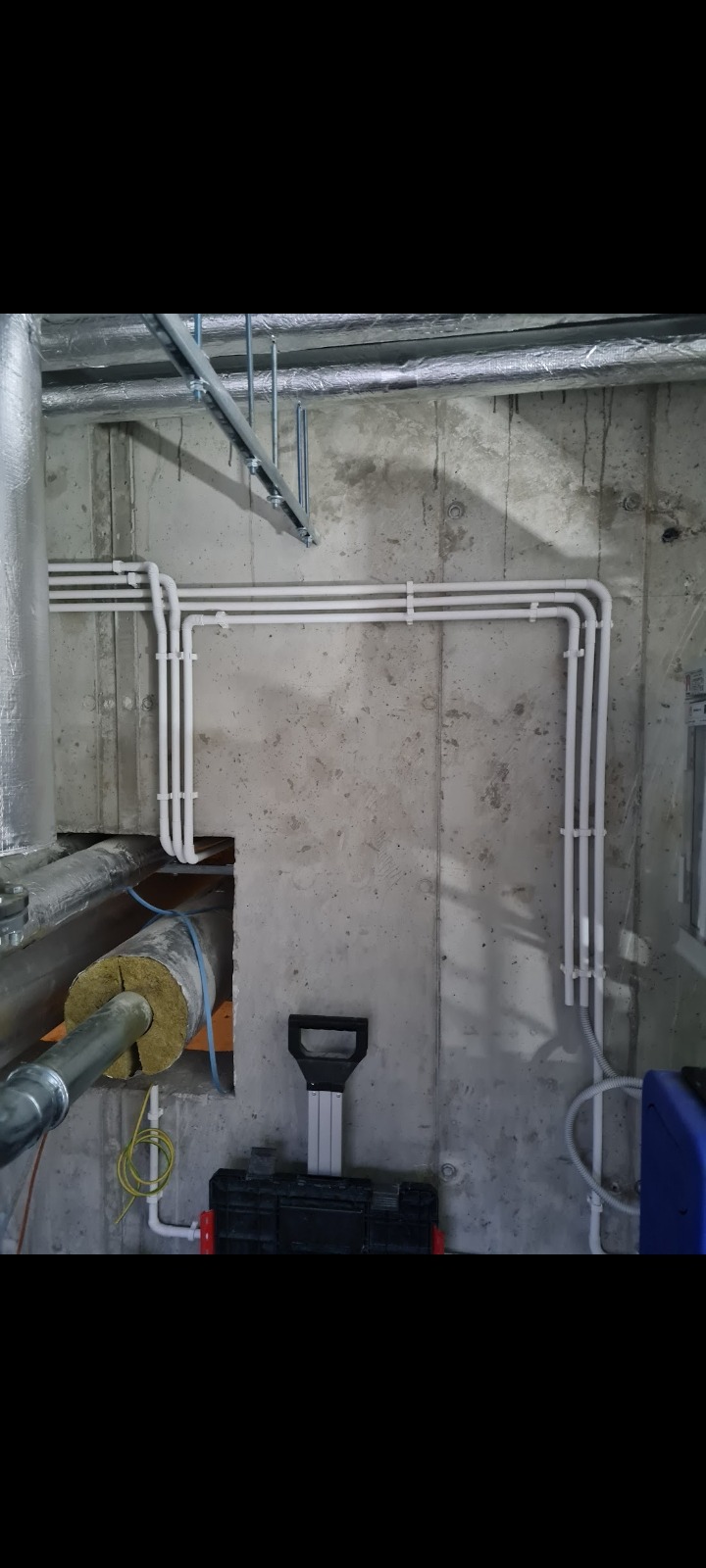 Białe rury poprowadzone wzdłuż surowej, betonowej ściany, widoczne przewody w izolacji i skrzynka narzędziowa na podłodze.