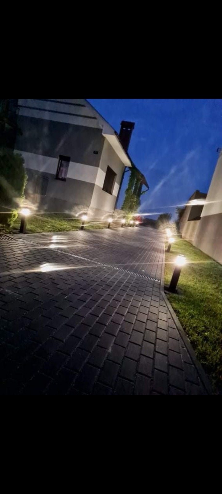 Oświetlona ścieżka z kostki brukowej prowadząca do domu w wieczornym oświetleniu, z lampami ogrodowymi po obu stronach.