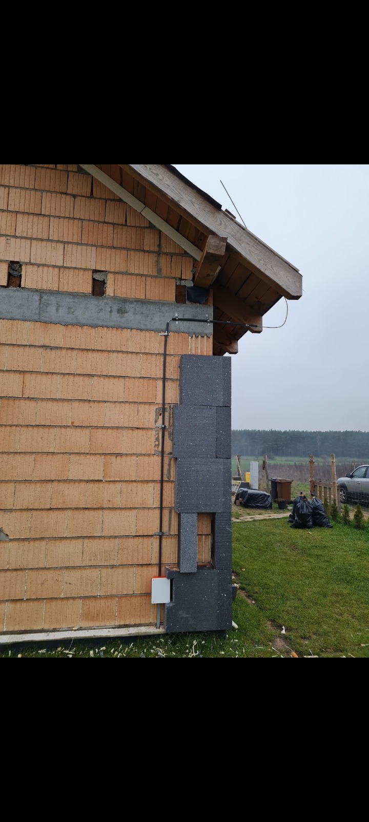 Fragment ściany budynku w trakcie ocieplania: widoczny mur z cegły, częściowo przykryty szarym styropianem, z zamontowaną puszką elektryczną i przewodami na zewnątrz.