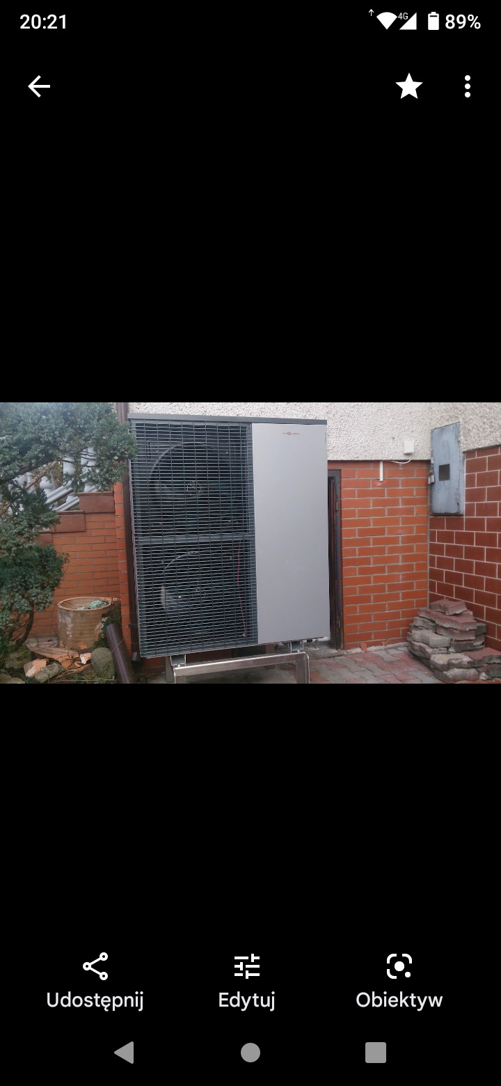 Szara pompa ciepła marki Viessmann zamontowana na metalowej konstrukcji przy ścianie budynku z czerwonej cegły, widoczny fragment ogrodu z roślinnością.