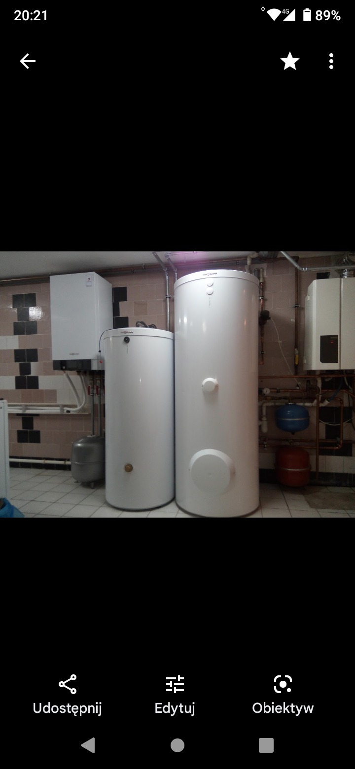 Dwa białe zbiorniki akumulacyjne ciepłej wody, jeden mniejszy i jeden większy, zainstalowane w kotłowni z widocznymi rurami, zaworami i innymi elementami instalacji. W tle widoczny piec grzewczy...