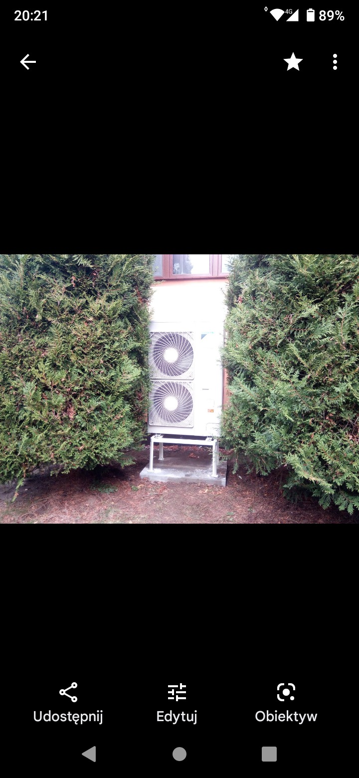 Zewnętrzna jednostka pompy ciepła Daikin zamontowana na betonowym fundamencie, otoczona tujami, widoczna z przodu budynku z oknem na górze.