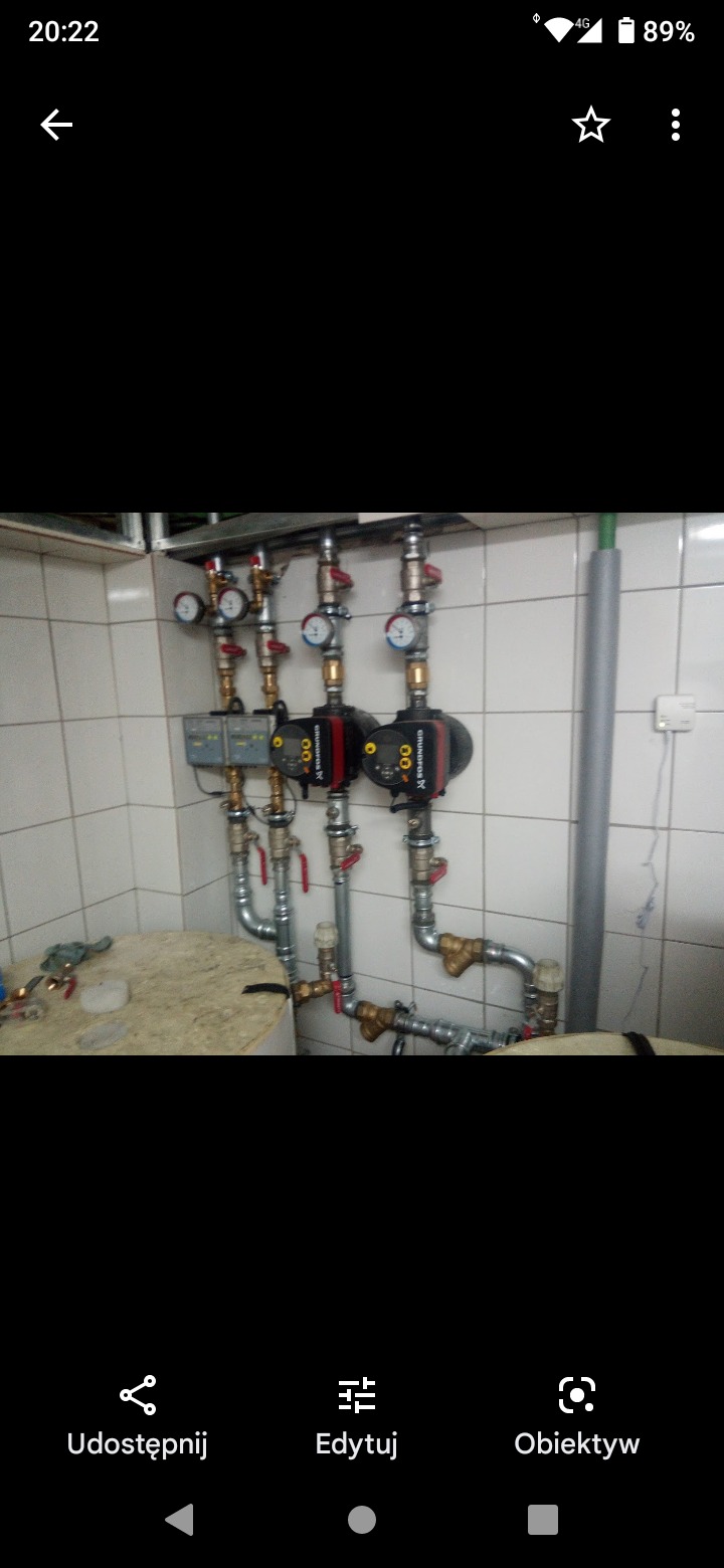 Dwie pompy obiegowe Grundfos zamontowane na ścianie z glazury, połączone rurami i manometrami, z widocznym fragmentem blatu roboczego i narzędziami w tle.