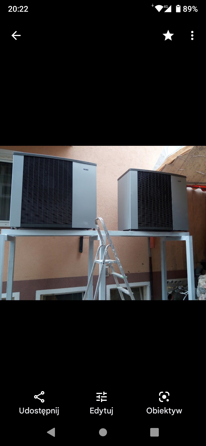 Dwie szare jednostki zewnętrzne na metalowej konstrukcji, z aluminiową drabiną opartą o konstrukcję, na tle beżowej ściany budynku.