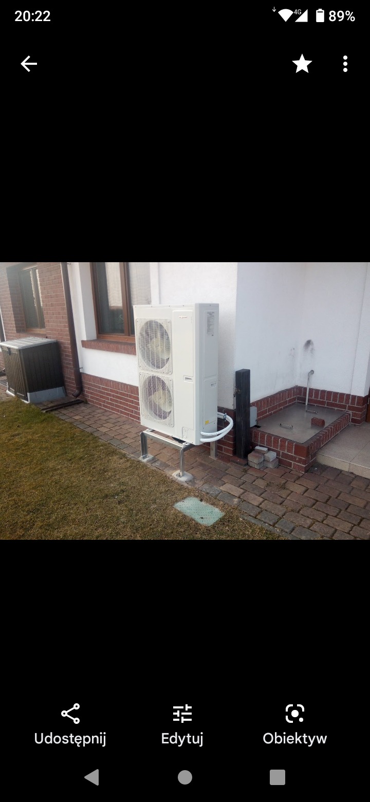 Zewnętrzna jednostka klimatyzacyjna typu split, zamontowana na metalowym stelażu przy ścianie domu z ceglaną podmurówką i trawnikiem. Widoczne rury instalacyjne.