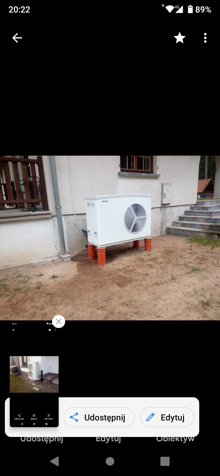 Biała jednostka zewnętrzna pompy ciepła na pomarańczowych rurach, ustawiona przy ścianie budynku z widocznym oknem i schodami wejściowymi.