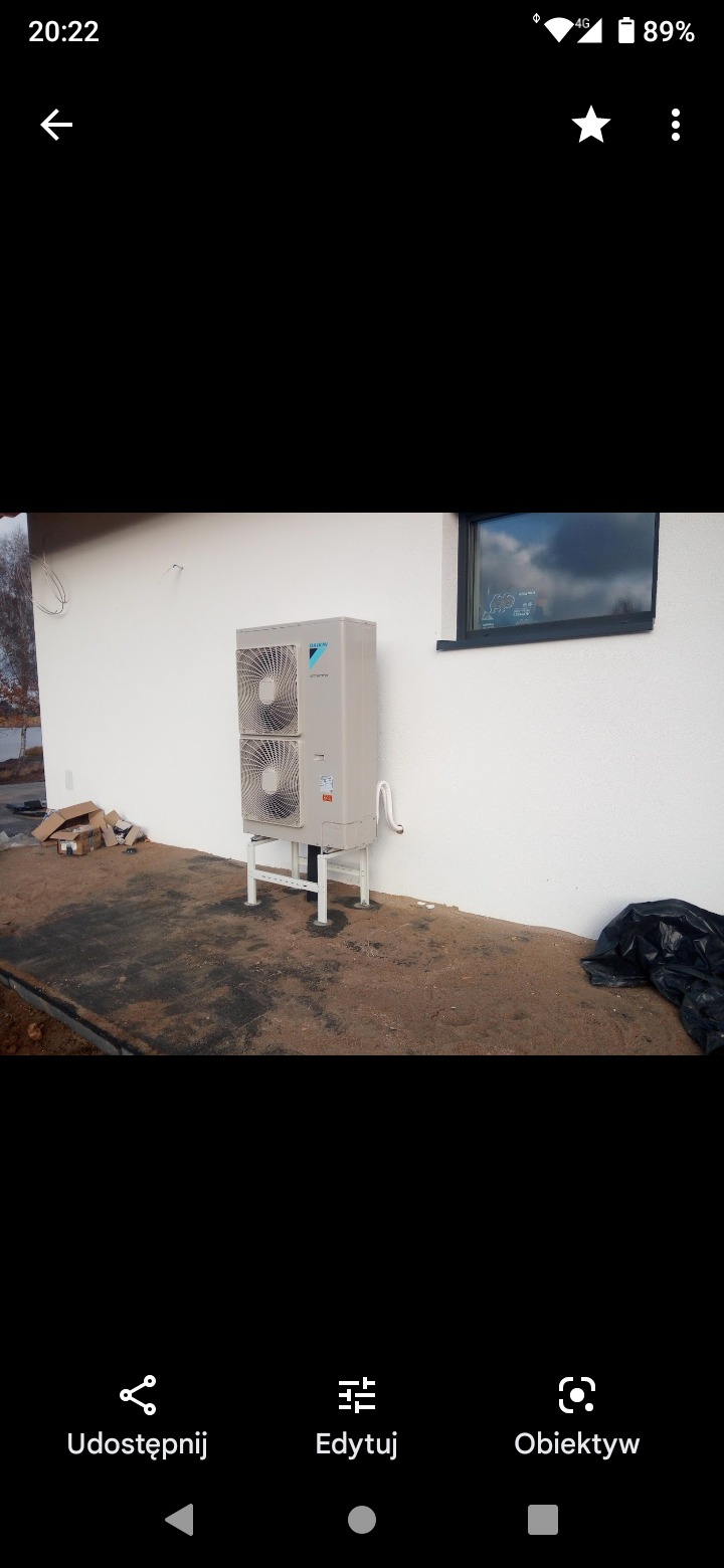 Zewnętrzna jednostka klimatyzacyjna Daikin zamontowana na białych wspornikach przy białej ścianie budynku, z widocznymi przewodami i fragmentami materiałów budowlanych na ziemi.