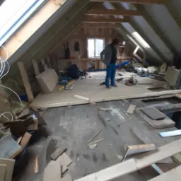 Poddasze w trakcie adaptacji: widoczne krokwie, okna dachowe, częściowo ułożona podłoga z płyt OSB, narzędzia i materiały budowlane, oraz sylwetka osoby w jeansach i kurtce.