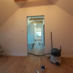 Poddasze po położeniu gładzi, widoczny fragment drewnianej belki stropowej, odkurzacz przemysłowy i pędzel malarski na podłodze z paneli.