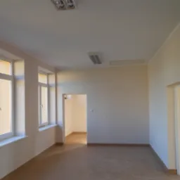 Puste, jasne pomieszczenie po remoncie z beżowymi ścianami, terakotą na podłodze i dwoma oknami z widokiem na zieleń. Widoczne drzwi do innego pokoju.