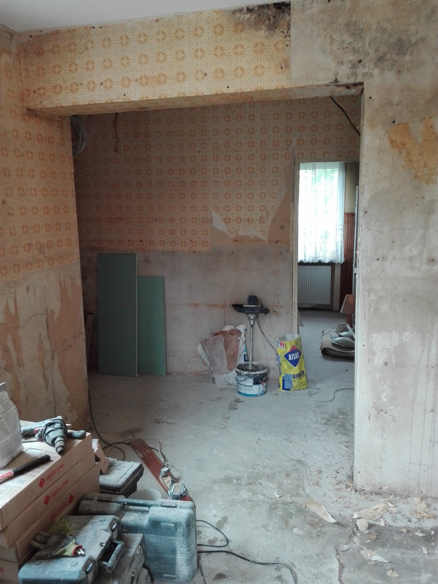 Pokój w trakcie remontu: zerwana tapeta z wzorem, surowe ściany, narzędzia i materiały budowlane, widoczne ślady wilgoci i grzyba na ścianie.