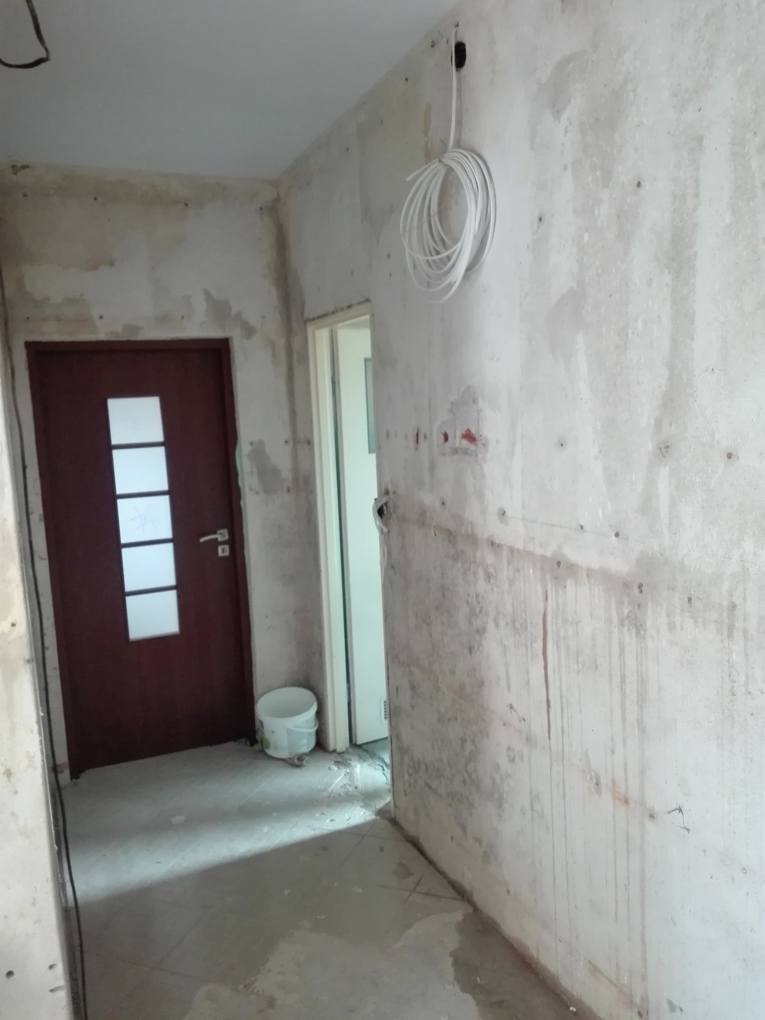 Korytarz w trakcie remontu: surowe ściany z widocznymi śladami po instalacjach, zwisający kabel elektryczny, brązowe drzwi z szybami i plamy na podłodze.