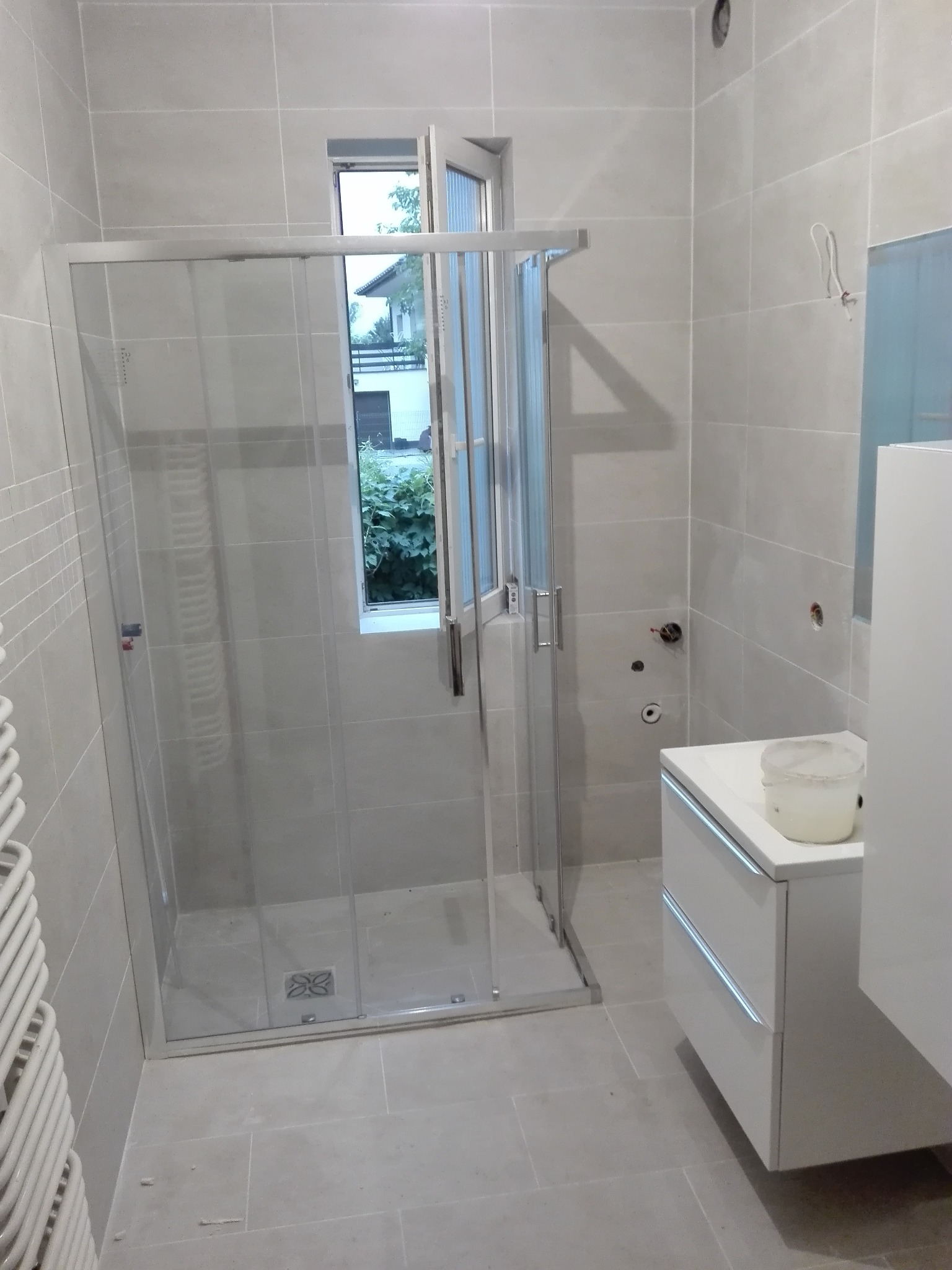 Jasna łazienka w trakcie prac wykończeniowych: kabina prysznicowa z przesuwnymi drzwiami, biała szafka umywalkowa z pojemnikiem na wierzchu, okno z widokiem na zieleń, płytki na ścianach i podłodze...