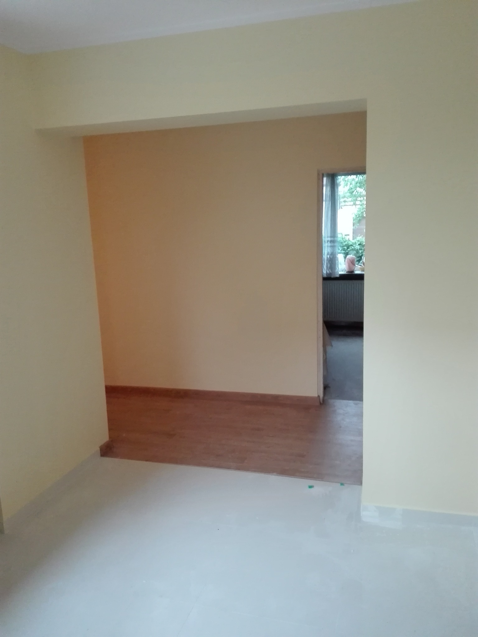 Pomalowane na beżowo wnętrze z fragmentem ułożonego parkietu, widok przez otwór drzwiowy na pokój z grzejnikiem i oknem.