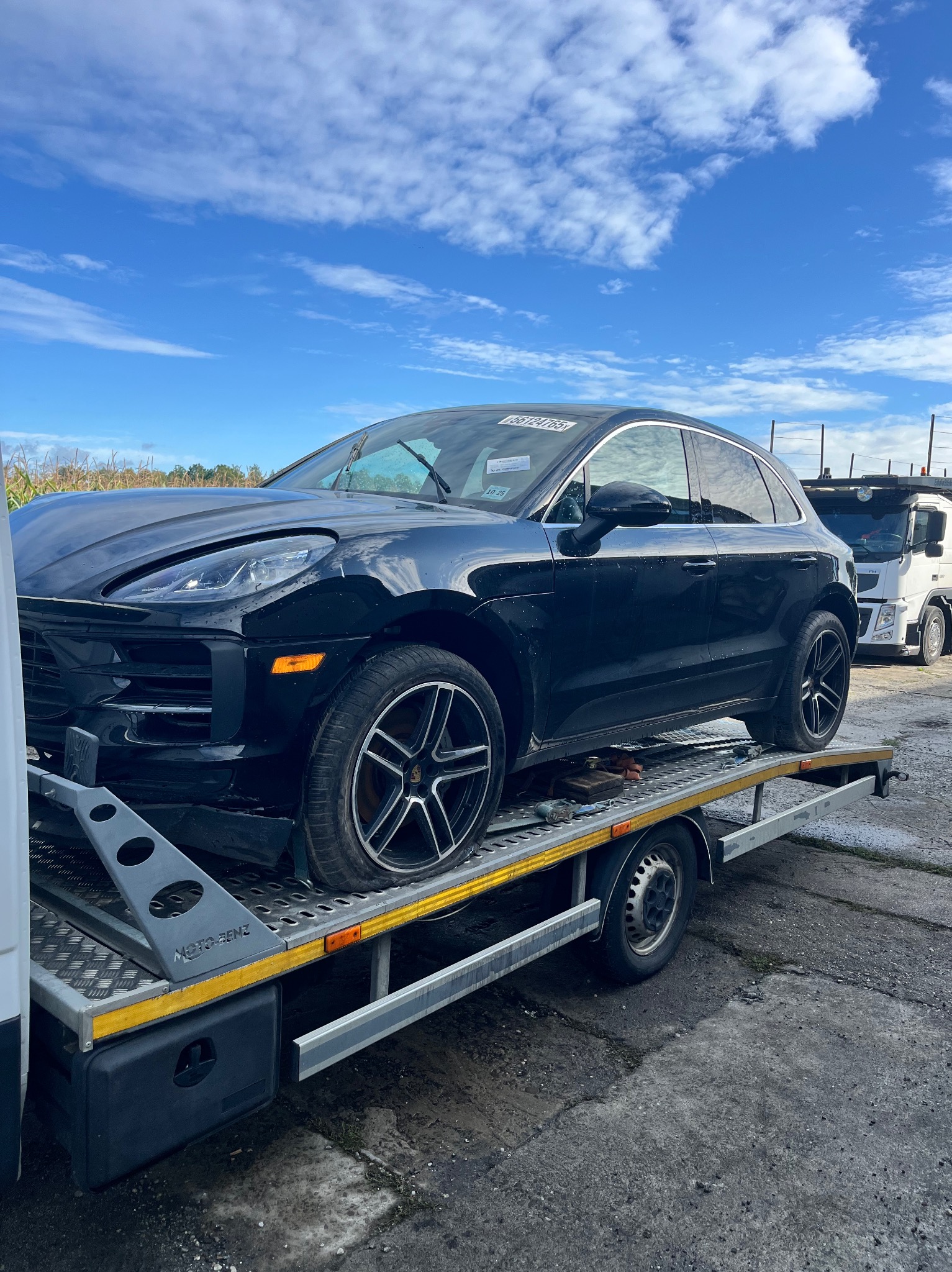 Czarny Porsche Macan z uszkodzonym przodem na platformie lawety Moto-Senz, na tle błękitnego nieba z chmurami i zaparkowanej lawety. Widoczne narzędzia.