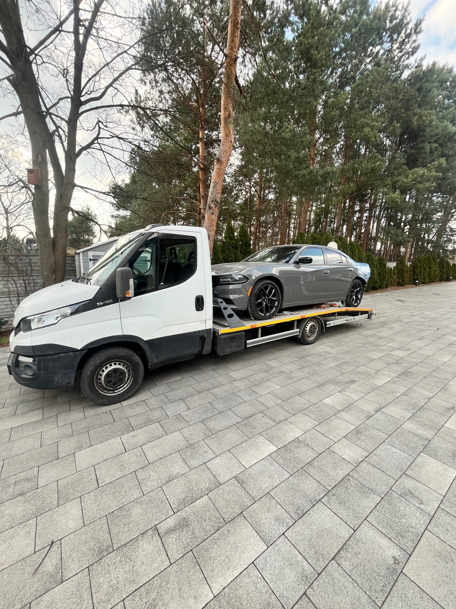 Szara osobówka na żółtej lawecie Iveco 35-130, na tle drzew i kostki brukowej. Widok z boku, lekko z dołu. Auto zabezpieczone pasami. Dzień, pochmurno.