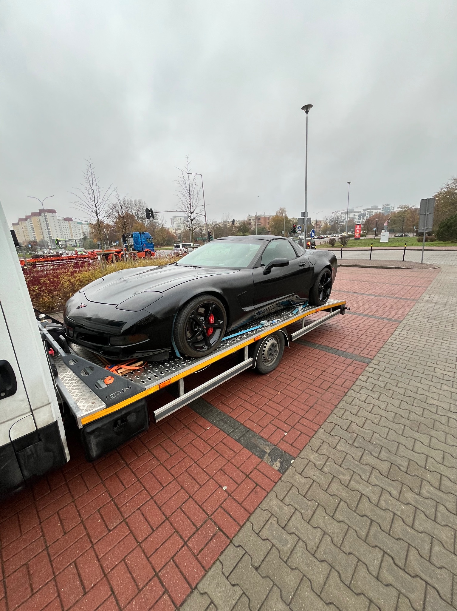 Czarny sportowy samochód Corvette na srebrnej lawecie, zabezpieczony pasami transportowymi, na tle miejskiego krajobrazu w pochmurny dzień.