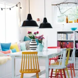 Jasne, skandynawskie wnętrze jadalni z białym stołem, kolorowymi krzesłami, poduszkami w oknie i czarnymi lampami wiszącymi.