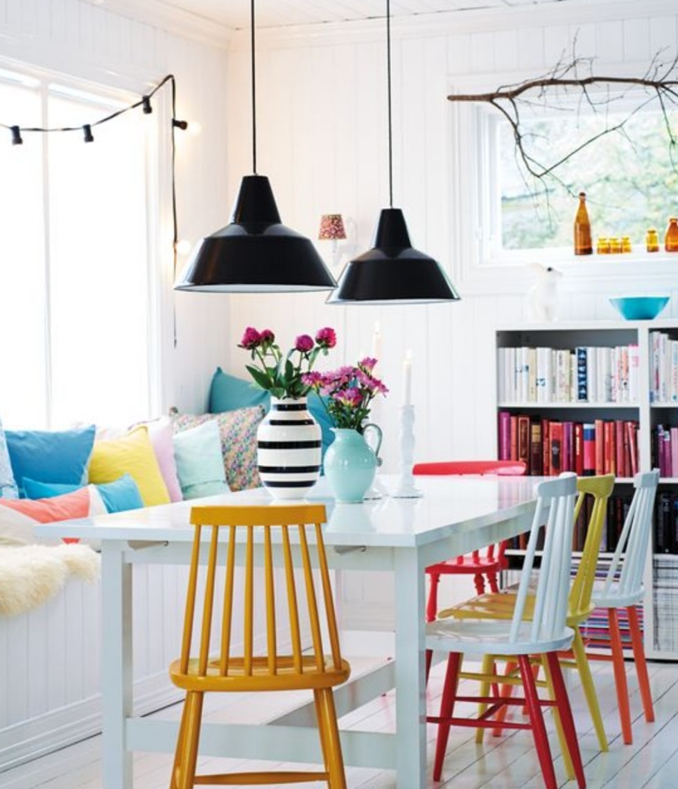 Jasne, skandynawskie wnętrze jadalni z białym stołem, kolorowymi krzesłami, poduszkami w oknie i czarnymi lampami wiszącymi.