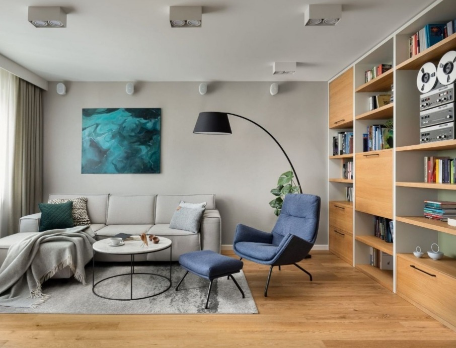 Jasne, nowocześnie urządzone wnętrze salonu z drewnianą podłogą, szarą sofą, błękitnym fotelem i regałem z jasnego drewna zabudowanym na ścianie.
