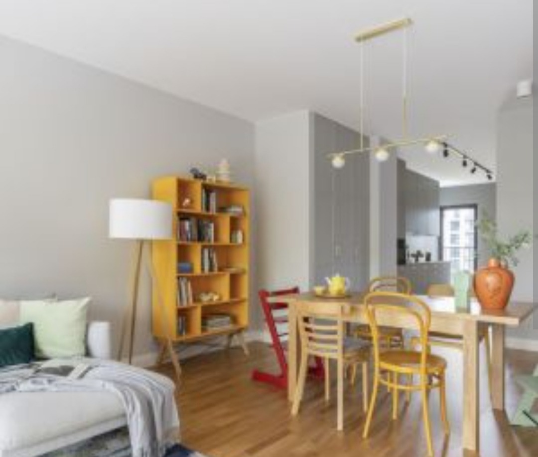 Jasne wnętrze mieszkania z drewnianą podłogą, żółtym regałem, białym narożnikiem i drewnianym stołem z krzesłami w różnych kolorach, oświetlone wiszącą lampą.