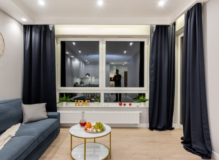 Nowoczesny salon z niebieską sofą, okrągłym stolikiem z owocami i ciemnymi zasłonami, widok na kuchnię w odbiciu okna.
