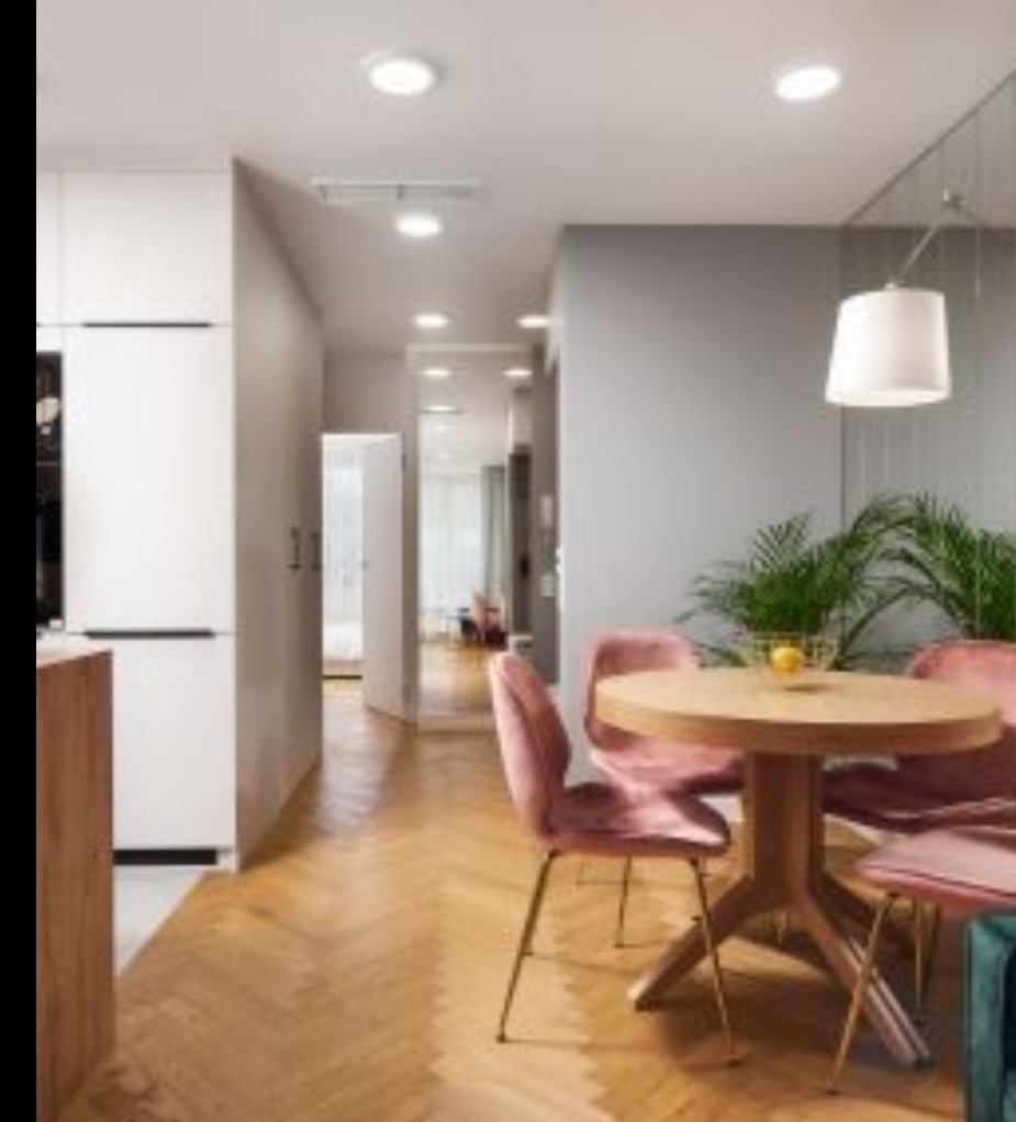 Jasne wnętrze mieszkania z drewnianą podłogą w jodełkę, okrągłym stołem z czterema różowymi krzesłami welurowymi i roślinami doniczkowymi na tle szarej ściany.