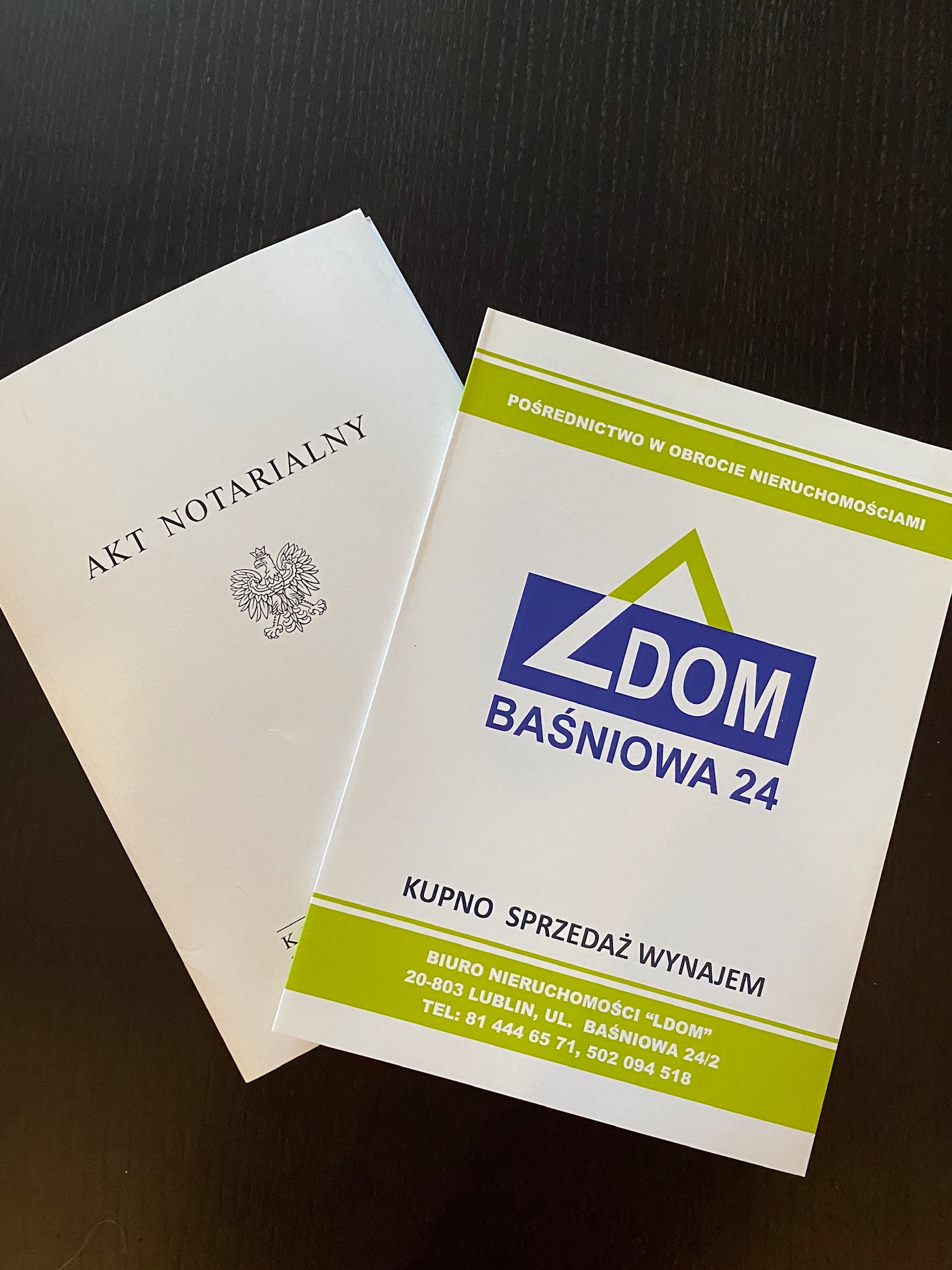 Dokument 'Akt Notarialny' obok ulotki biura nieruchomości 'LDOM' z Lublina, oferującego pośrednictwo, kupno, sprzedaż i wynajem, leżące na ciemnym blacie.