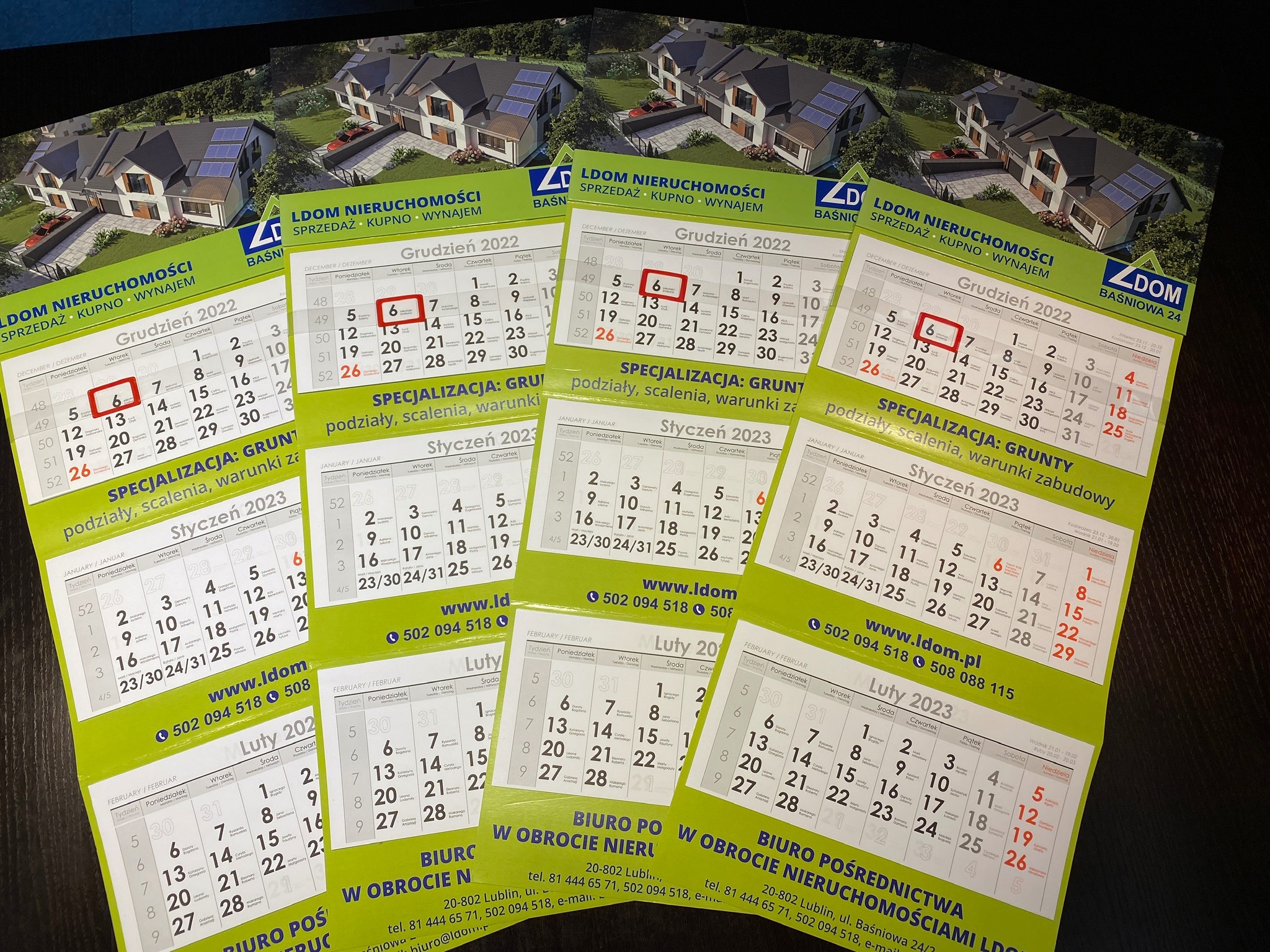 Stos kalendarzy biurkowych agencji nieruchomości LDOM, z widocznym logo, zdjęciem domu i kalendarium na grudzień 2022 i styczeń 2023, leżące na ciemnym podłożu.