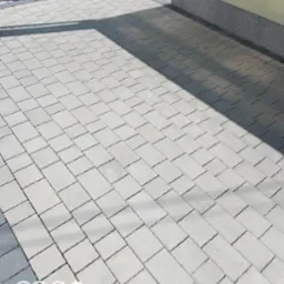 Ułożona szara kostka brukowa przed budynkiem z widocznym cieniem, donica z kwiatami przy ścianie budynku.