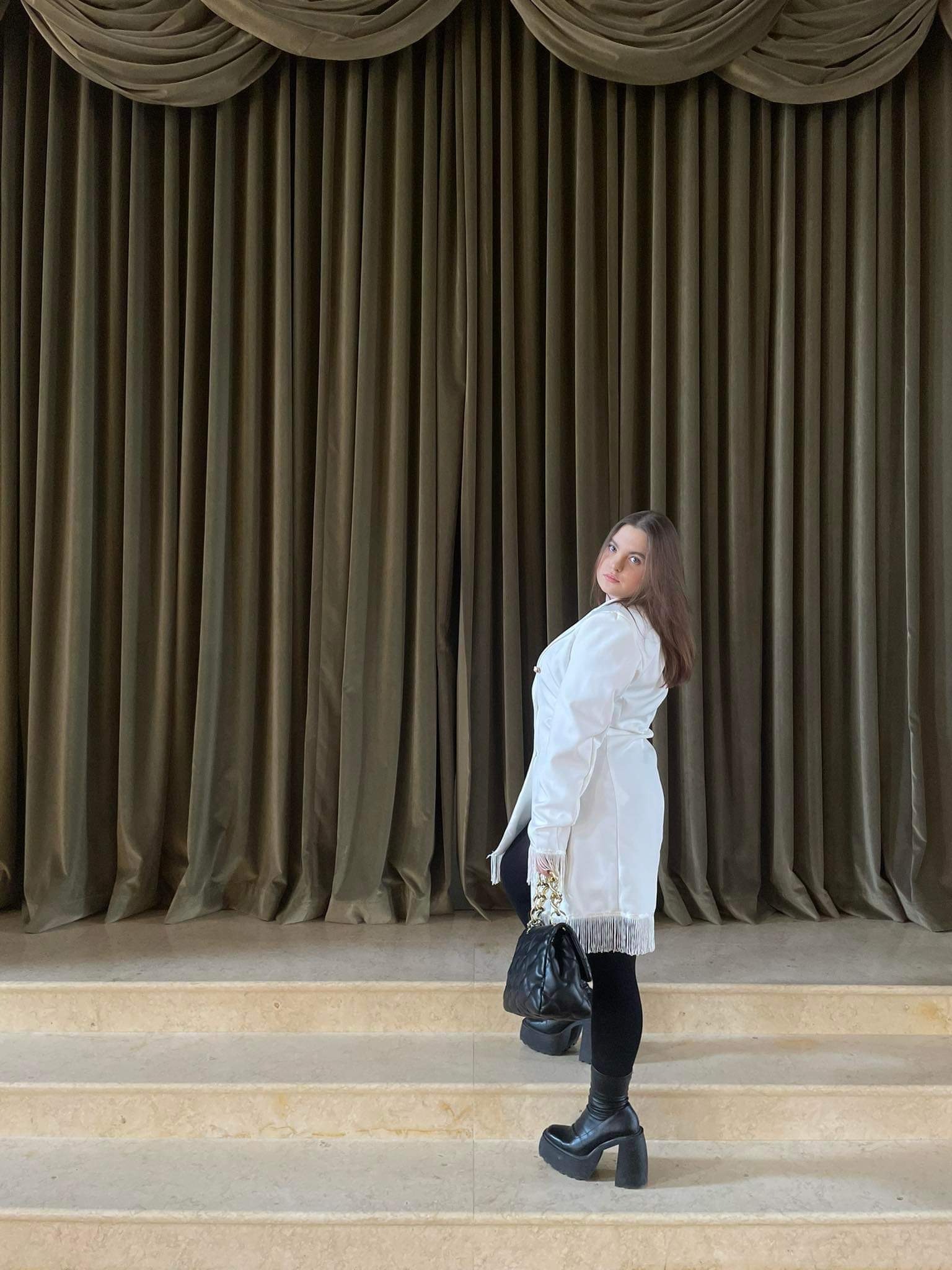 Kobieta w białej sukience z frędzlami, czarnych legginsach i botkach na platformie, pozująca na tle drapowanych, oliwkowych zasłon i marmurowych schodów w Łodzi, trzymająca czarną torebkę...