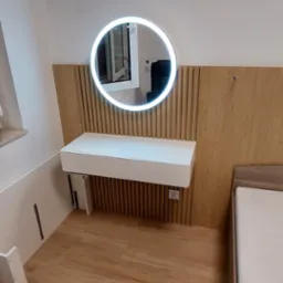 Nowoczesna sypialnia z toaletką z podświetlanym okrągłym lustrem i drewnianym panelem ściennym, obok łóżko z jasną pościelą, widoczne gniazdko elektryczne.