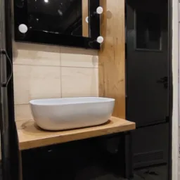 Nowoczesna łazienka z umywalką nablatową, lustrem podświetlanym żarówkami, drewnianym blatem i czarnymi szafkami na wysoki połysk.