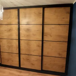 Duża szafa wnękowa z przesuwnymi drzwiami o drewnianej fakturze, podzielona na kwadratowe panele z czarnymi ramami, oświetlona przez kryształowy żyrandol.