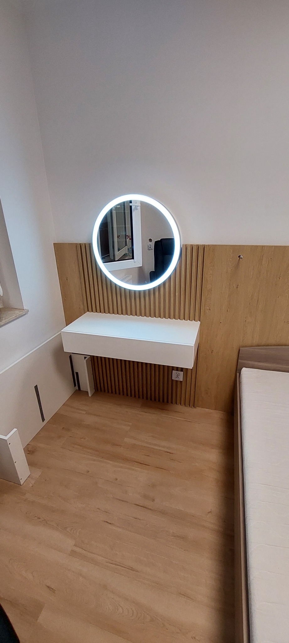 Nowoczesna sypialnia z toaletką z podświetlanym okrągłym lustrem i drewnianym panelem ściennym, obok łóżko z jasną pościelą, widoczne gniazdko elektryczne.