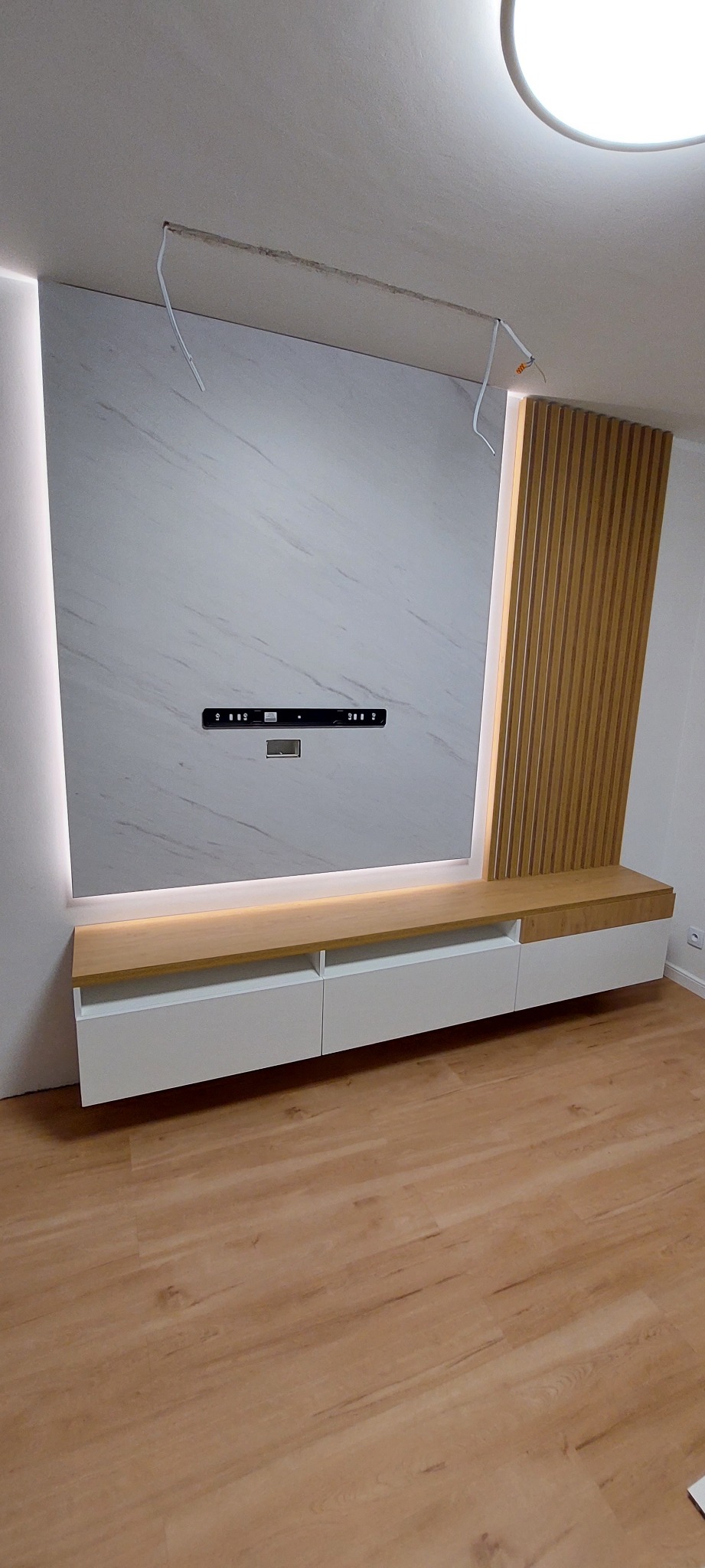 Nowoczesna ściana telewizyjna z marmurową okładziną, podświetleniem LED, drewnianymi lamelami i białą szafką RTV z drewnianym blatem, widoczne kable wystające z sufitu.