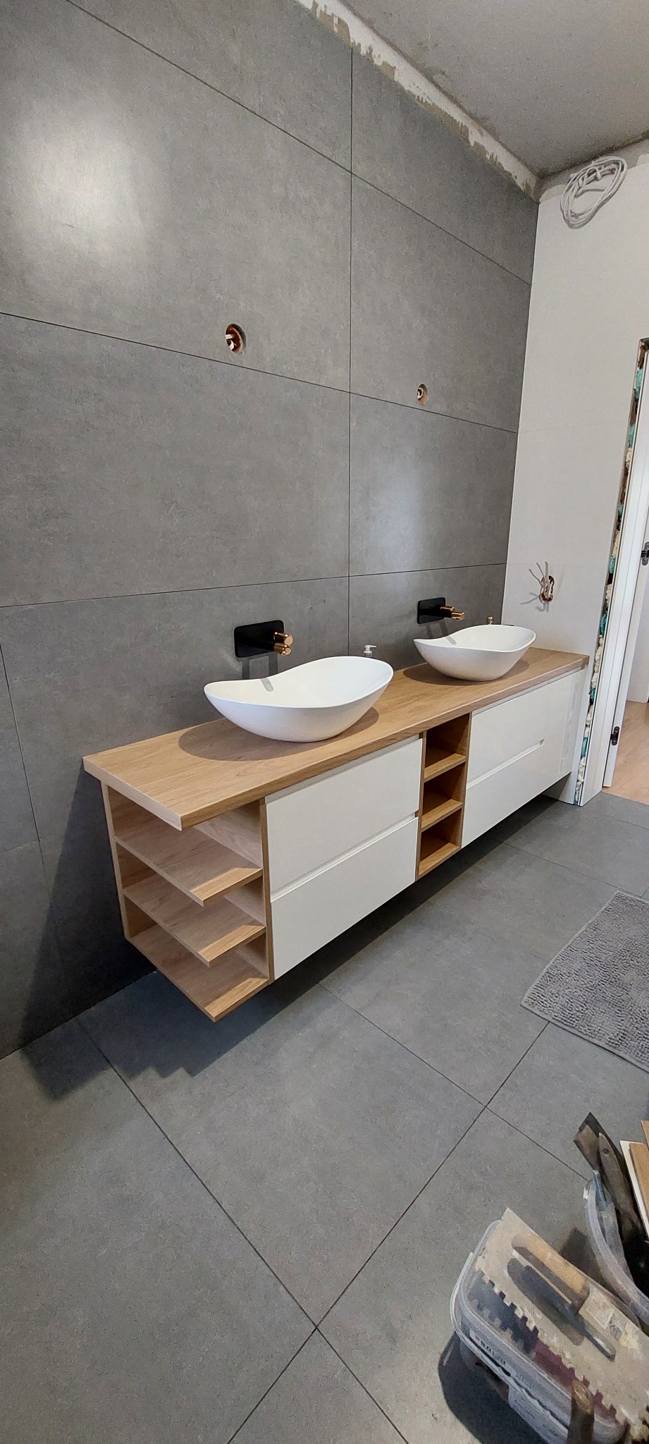 Łazienka w trakcie wykańczania: podwójna umywalka nablatowa z drewnianym blatem i białymi szafkami, szare płytki na ścianach z widocznymi instalacjami, na podłodze pojemnik z narzędziami.
