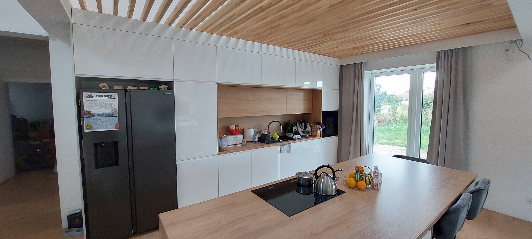 Nowoczesna kuchnia z białymi szafkami, drewnianymi akcentami i wyspą z płytą indukcyjną oraz dekoracyjnymi dyniami, oświetlona naturalnym światłem z okna.