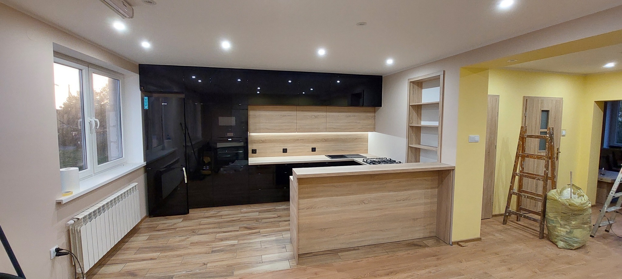 Nowoczesna kuchnia z czarnymi, błyszczącymi frontami i elementami drewnopodobnymi, oświetlona punktowo, z wyspą kuchenną i widocznymi elementami wykończeniowymi jak drabina i worki.